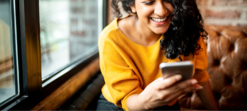 A young woman sitting by a window in a public space, smiling at what she is reading on her phone. Click to view article, Share Your Experience: Public Health Department Seeks Input on Community Health
