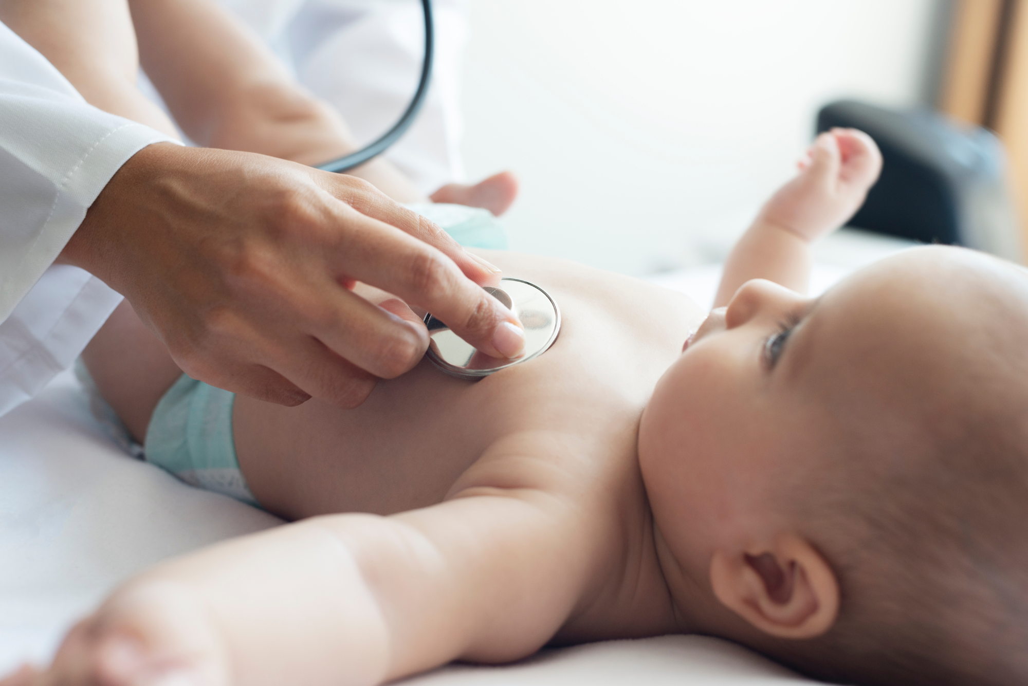Baby on an exam table having chest examined by a doctor using a stethoscope. Click to view article, Health Officials Urge Precautions as Pediatric RSV Hospitalizations Increase