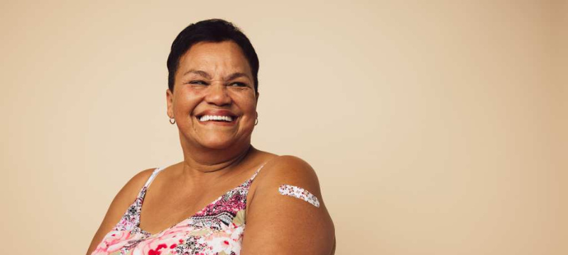 Woman in her 60s smiling wearing a flower-patterned blouse and a flower-patterned bandaid on her shoulder.
