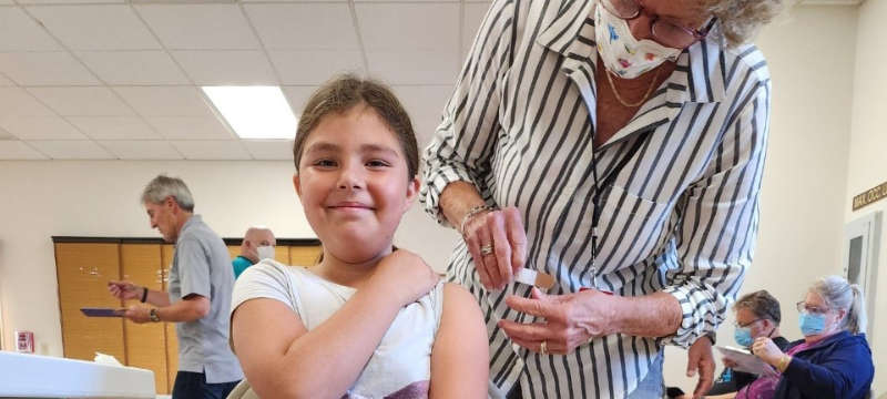 A young girl getting a bandaid placed on her shoulder be a nurse at a pop-up vaccine clinic.