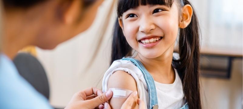 A girl has a white bandaid being placed on her shoulder by her pediatrician.