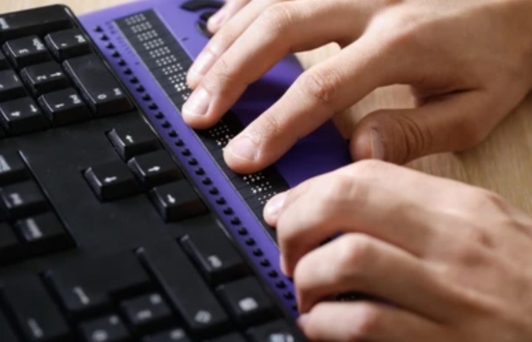 Two hands reading a refreshable Braille device, next to a black computer keyboard. Click to view article, Global Accessibility Awareness Day 2021