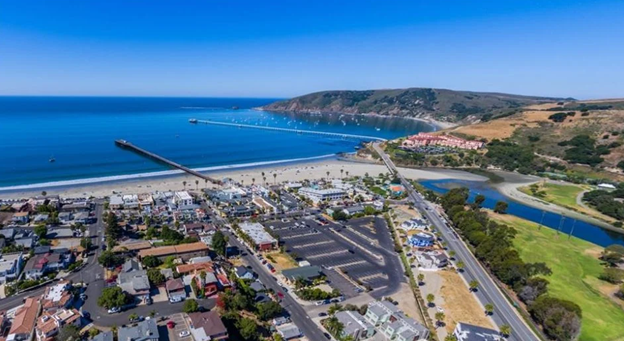 Aerial view of Avila Beach from inland looking out towards ocean.  The ocean, beach and pier are visible in the background and in the foreground several blocks of urban development are visible.