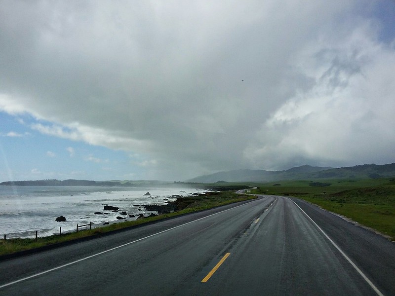 Road next to the ocean and green grass with clouds overhead