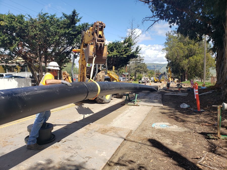 Man in safety vest and hardhat helps to guide large pipeline on street; green trees and blue skies in background Click to view article, Five SLO County Agencies Awarded Grant Funding for Water Projects
