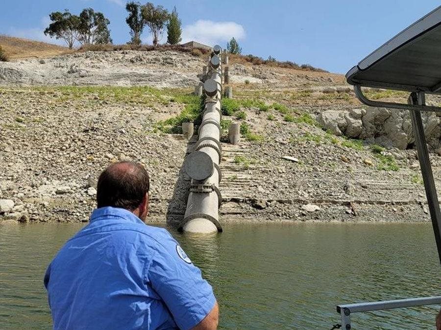 Employee in blue sits in front a body of water and a downhill pipeline  Click to view article, State Restrictions Lifted from Nacimiento Water Project