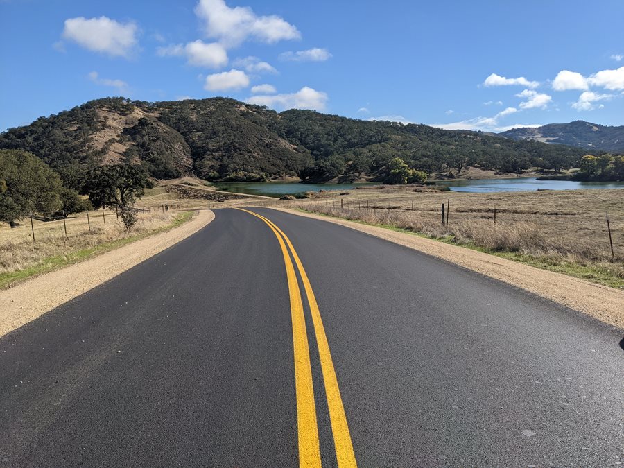 A newly-paved asphalt road stretches toward hills and a body of water in the background