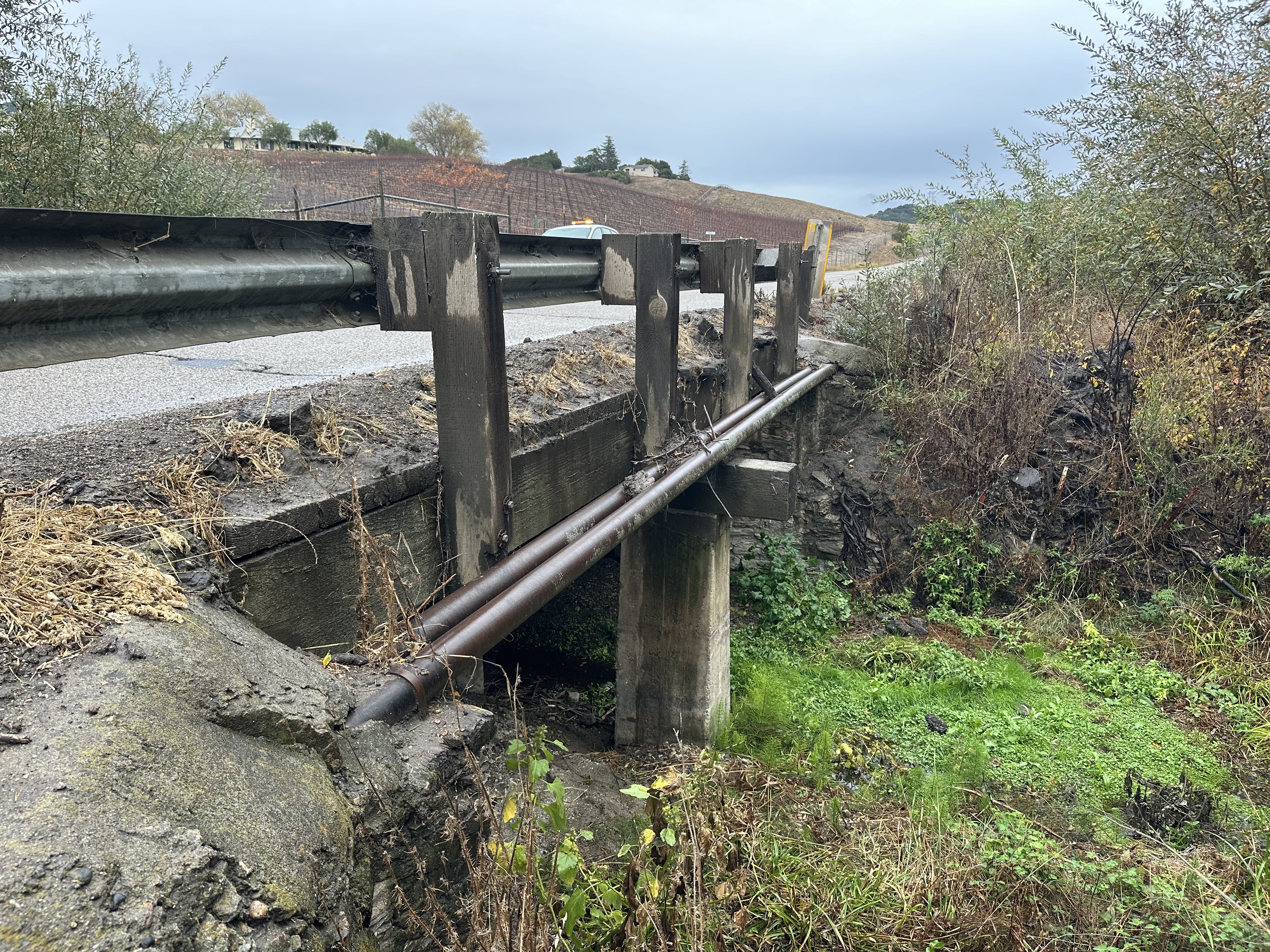 Wooden guardrail along a small rural bridge, with visible wear on the rail and concrete, surrounded by trees and vegetation