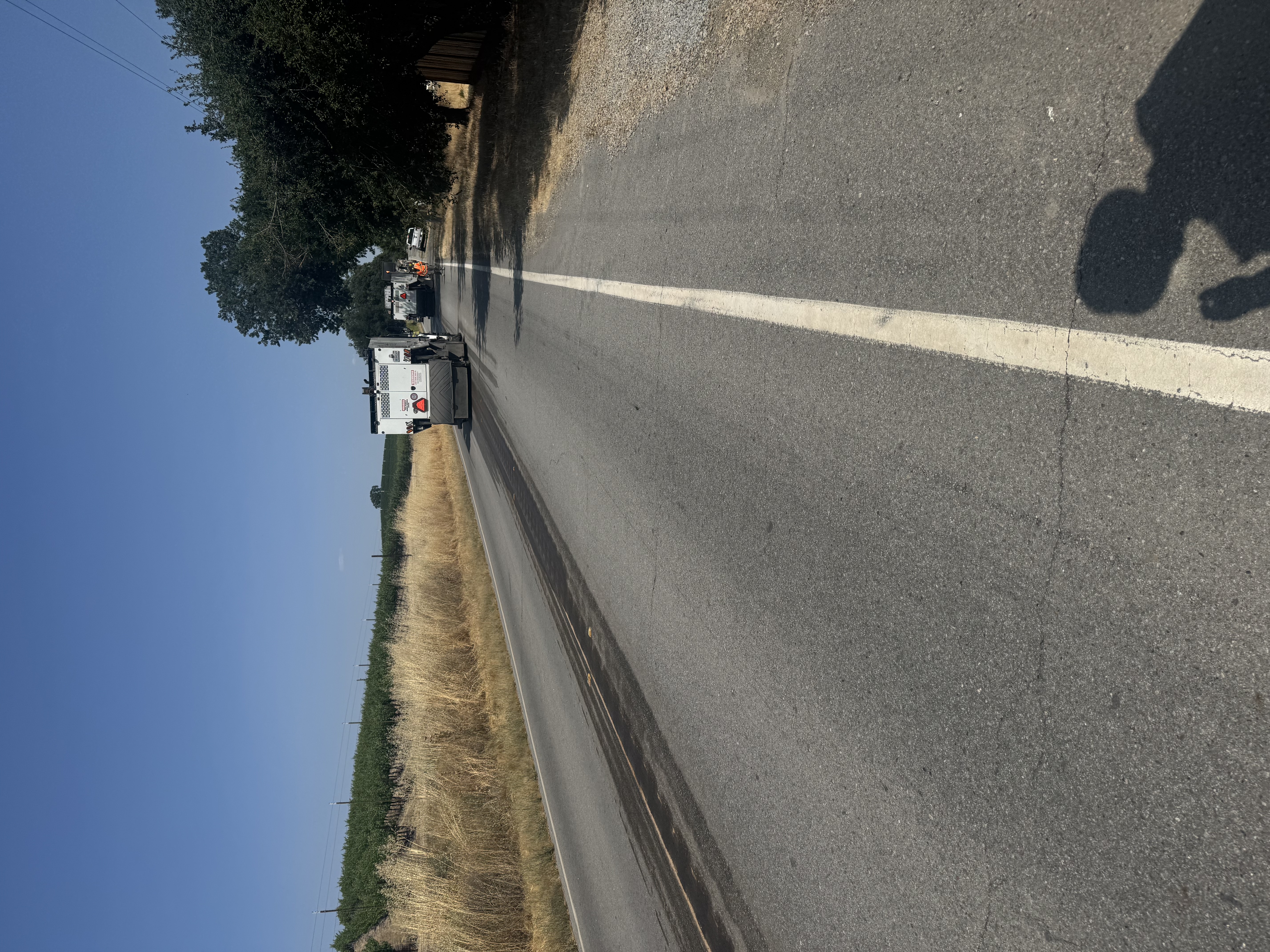 A large truck drives down the center of a paved road with tall grass and a vinyard in the background. Oak trees and workers stand to the side of the road.