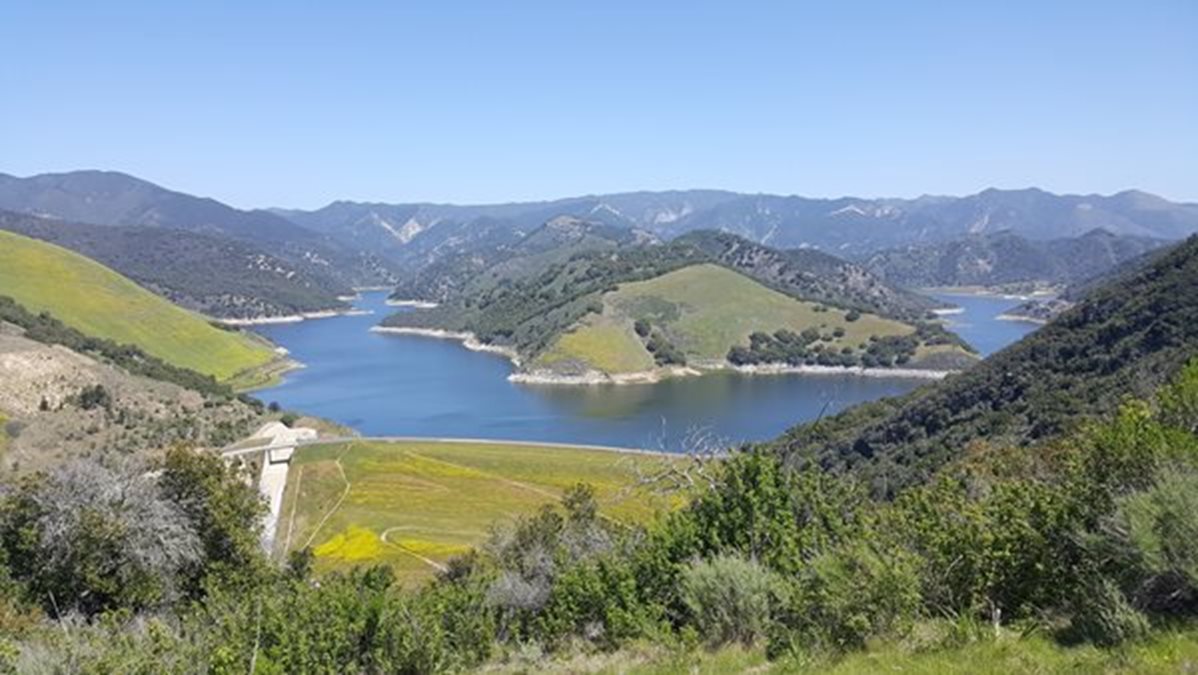 Panoramic view of a blue lake surrounded by rolling green hills and mountains under a clear blue sky.