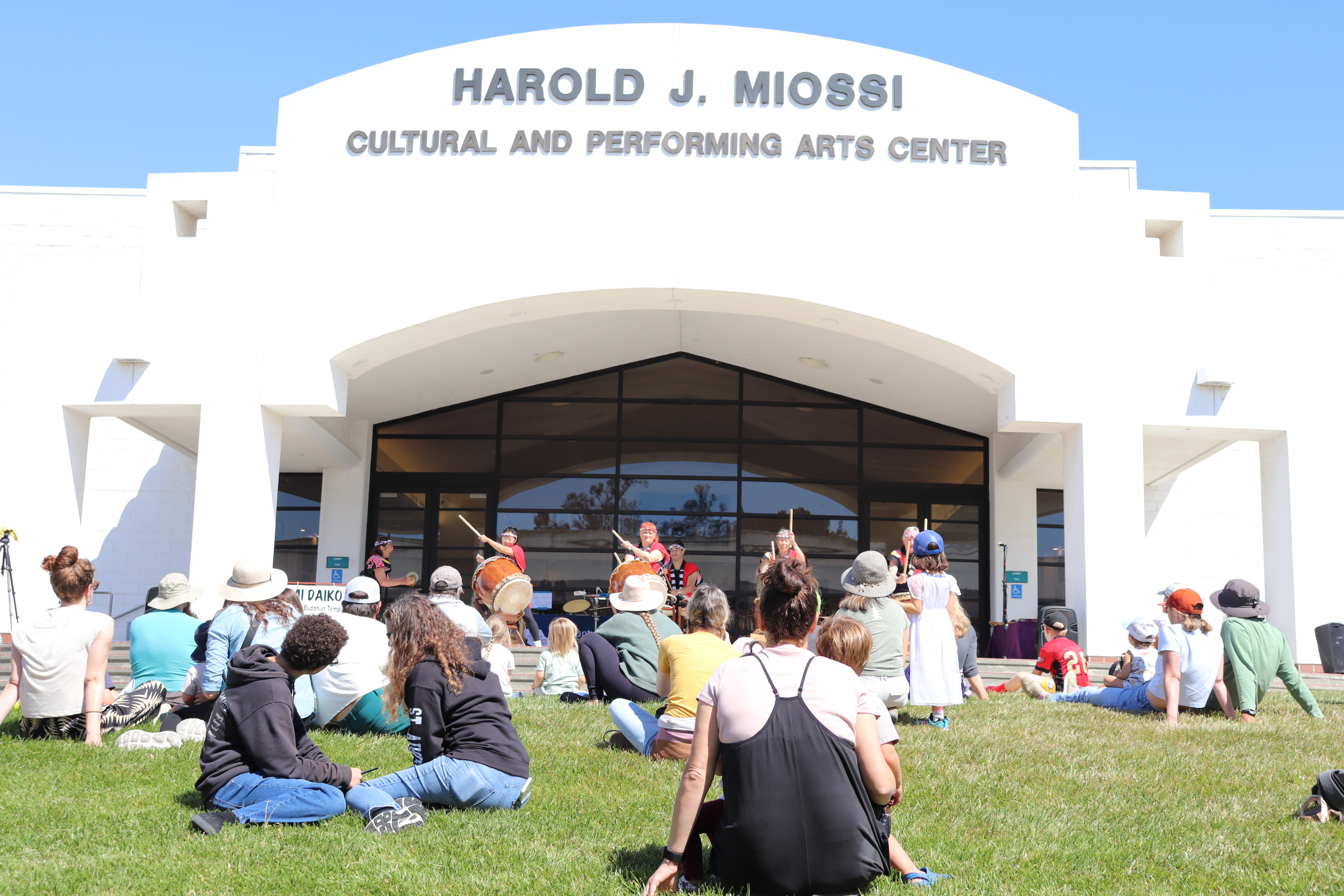 Families sitting in front of the grass in front of a building labeled Harold J. Miossi Cultural and Performing Arts Center