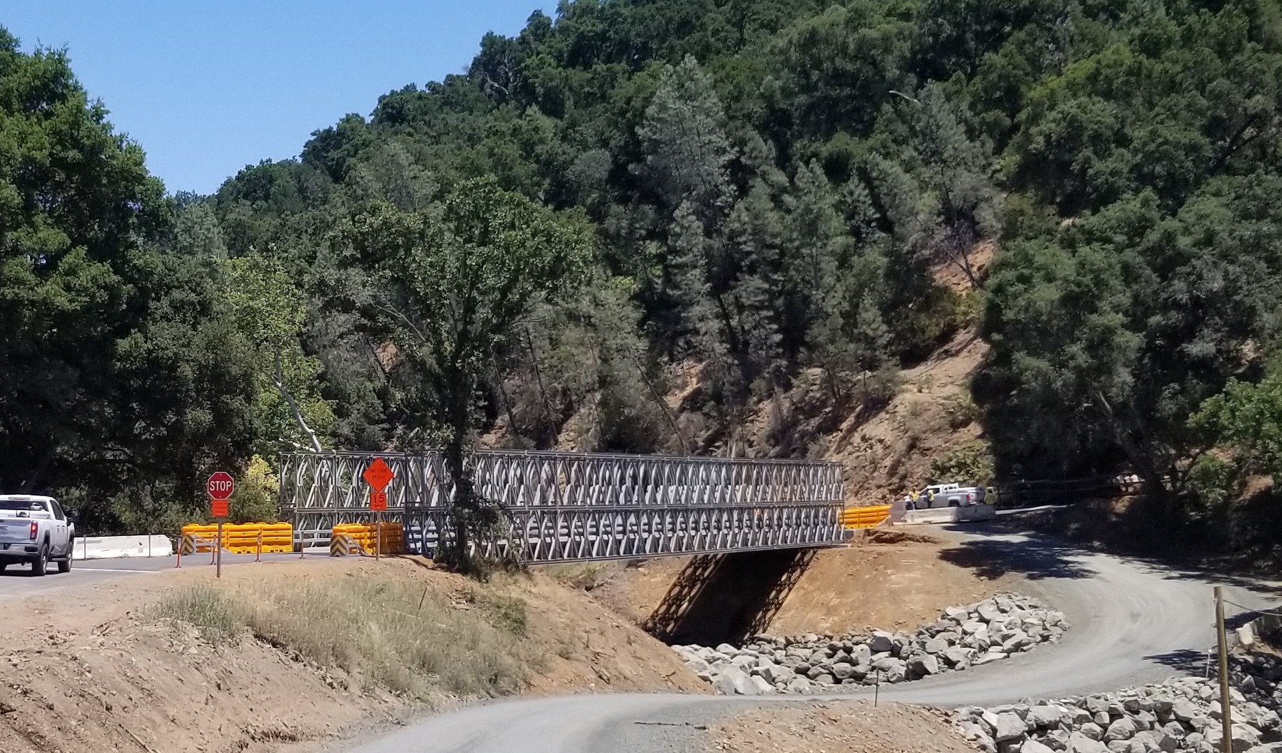 A bridge connects a paved road above a service road and rocky, dry, creekbed and hillsides with dense oak trees and blue skies above.