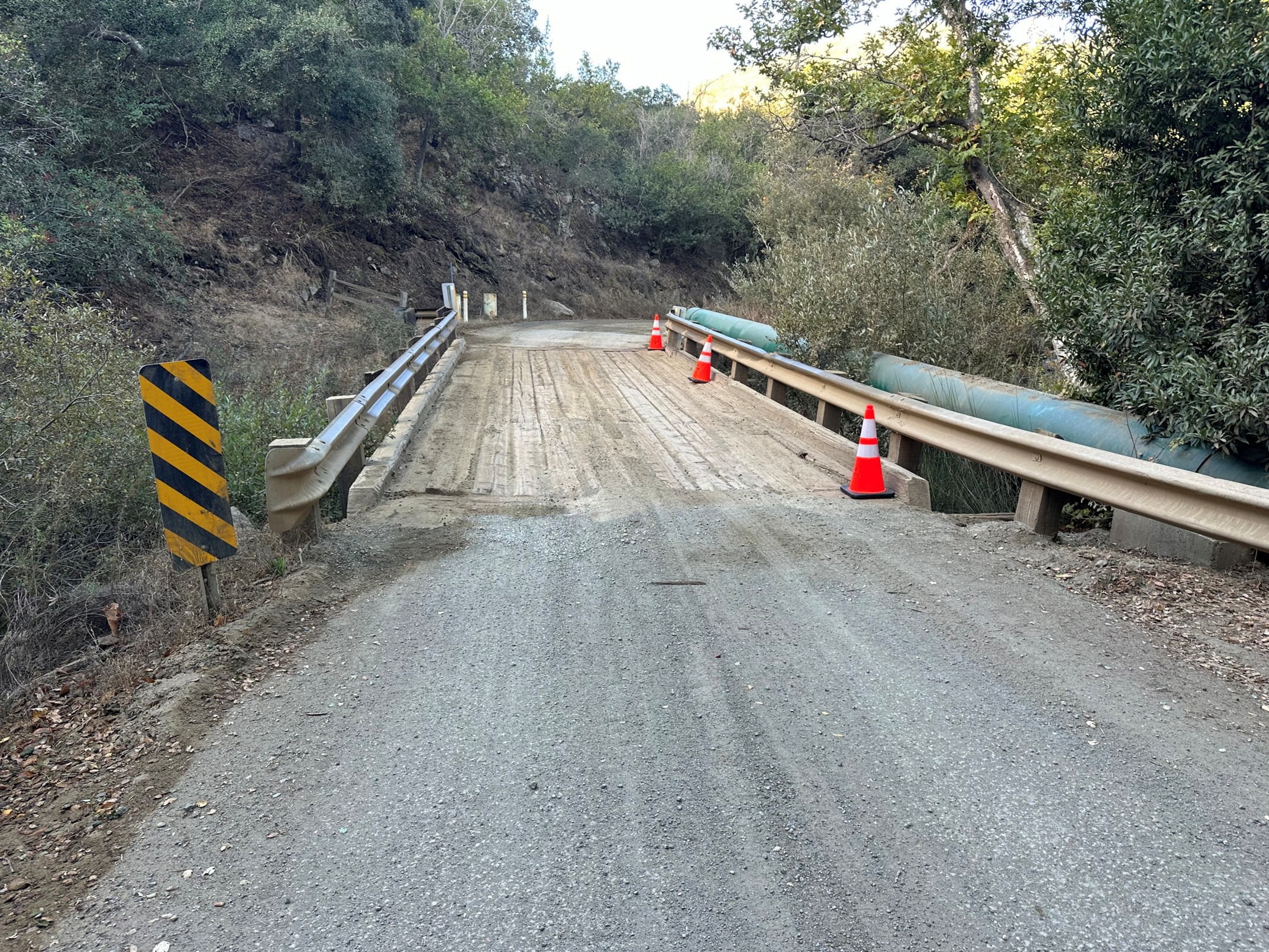 A dirt road leading onto a wooden bridge and a curving dirt road beyond. Three traffic cones are on the bridge, beside a hill with vegetation and above a ravine with vegetation.