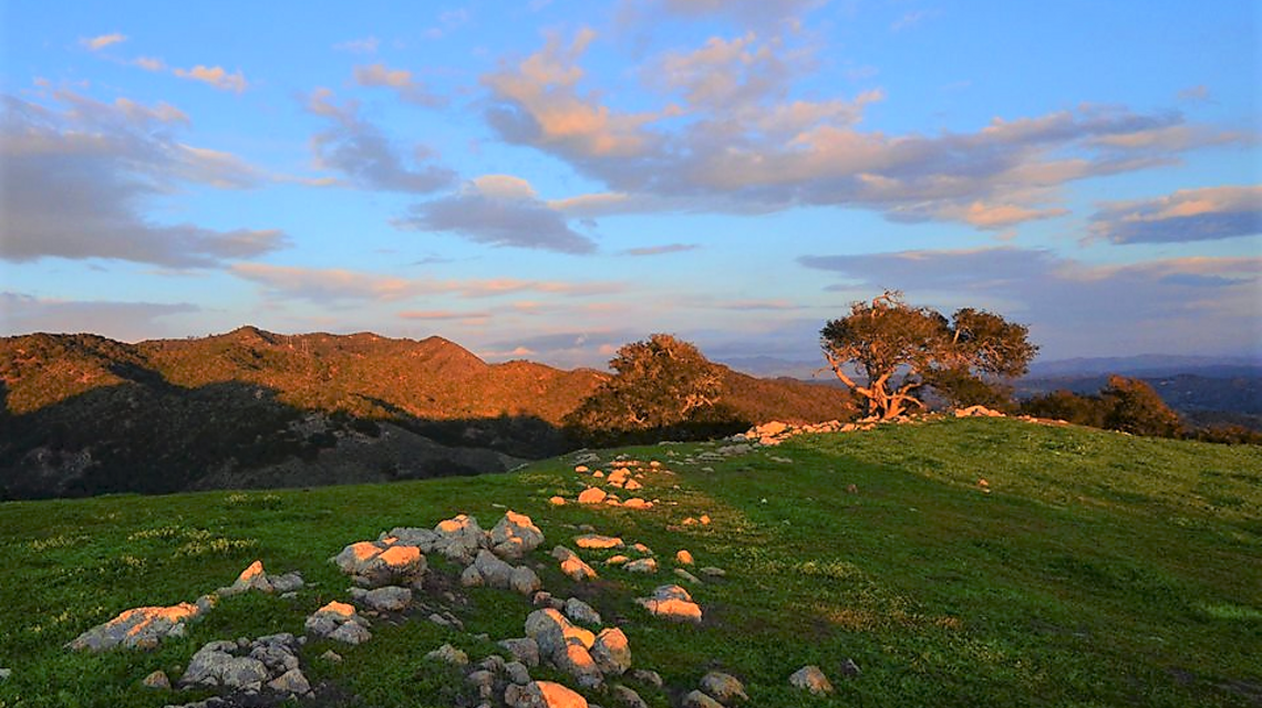 Image of a green hill with a rock trail. There is a blue sky with clouds and more mountains in the distance.