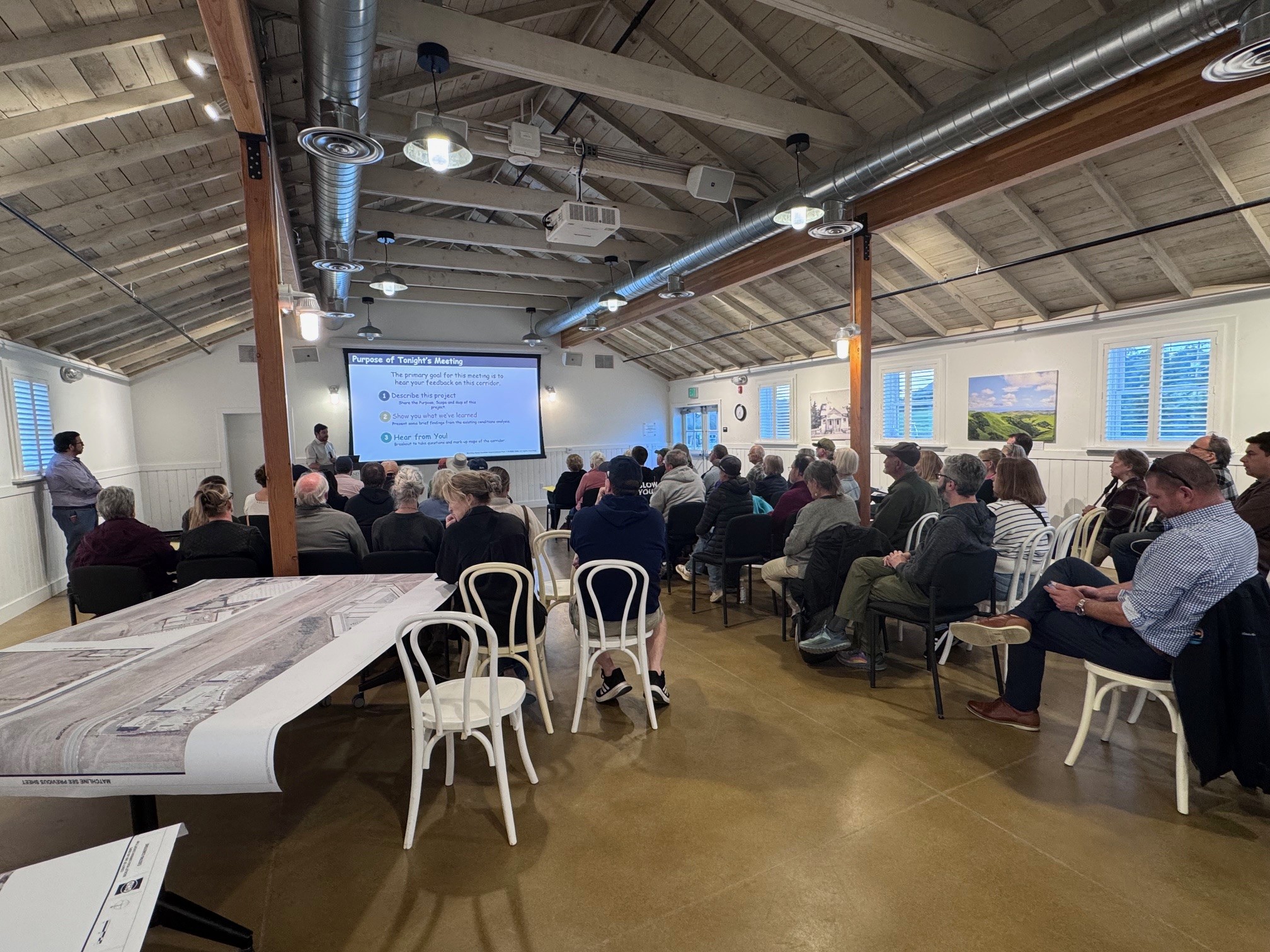 A group attends a meeting in a wood-beamed room, facing a presenter and projection screen.