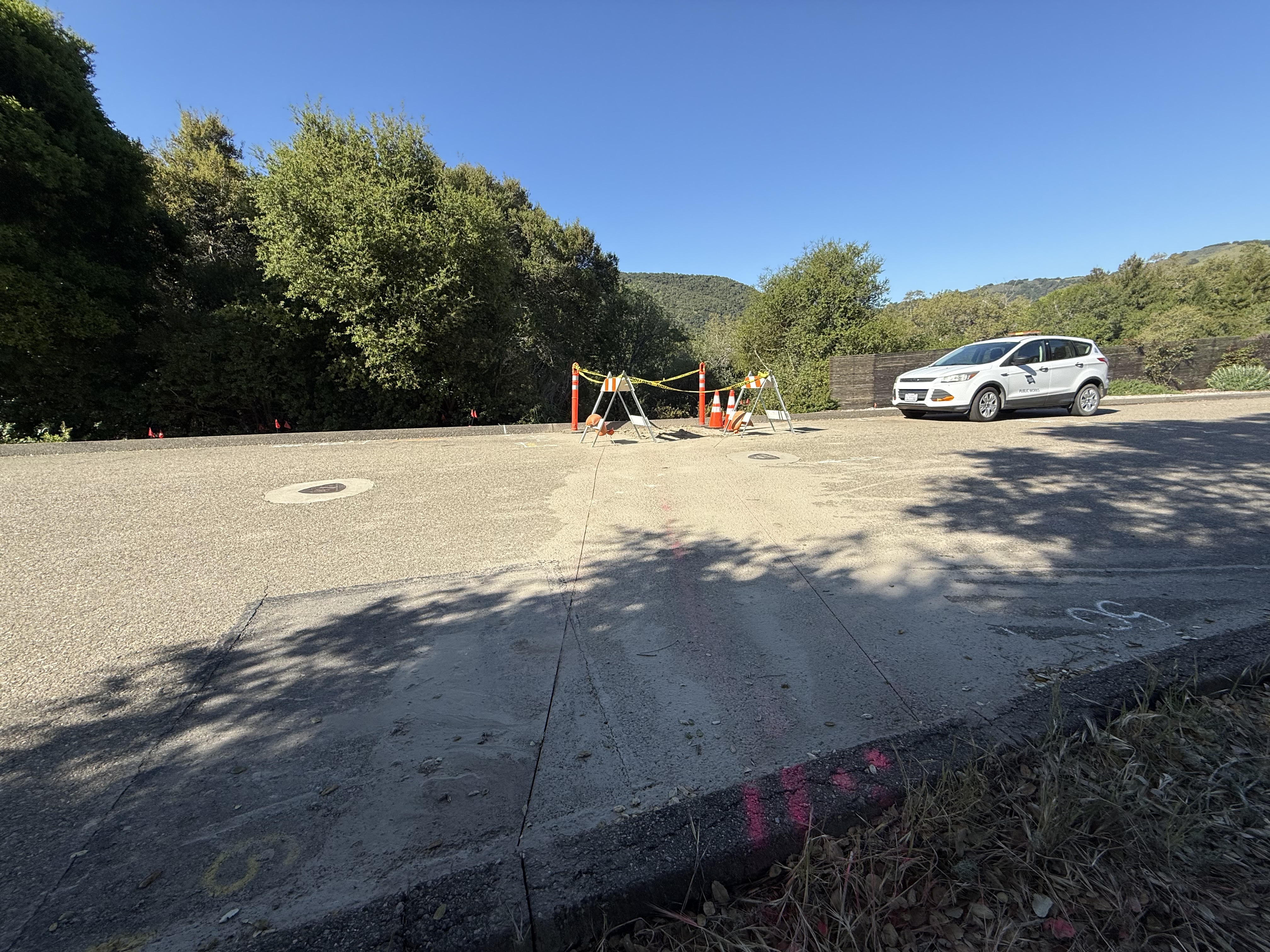 Roadside construction area with cones and caution tape, utility vehicle parked nearby, trees and hills in the background.