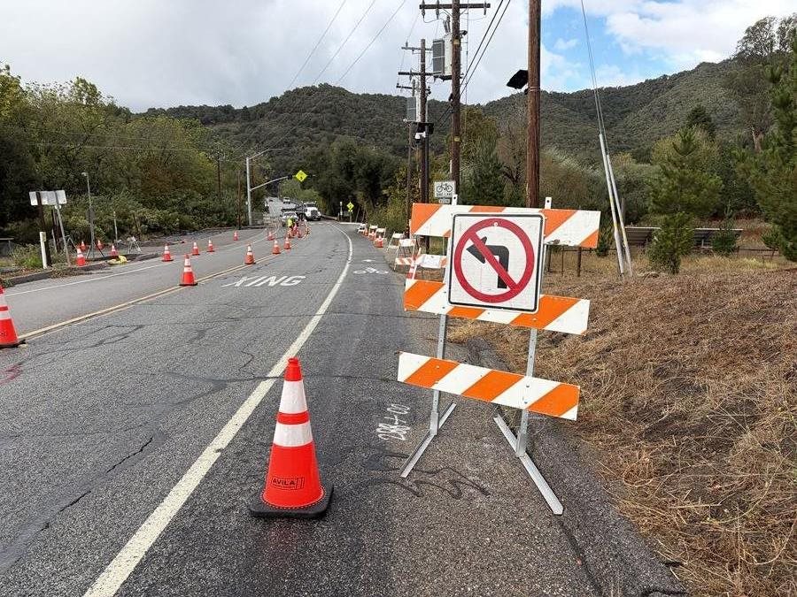 A two-lane road through wooded hills is partially closed for roadwork, with orange traffic cones directing vehicles and striped barricades displaying a no-left-turn symbol. Utility poles and overhead 