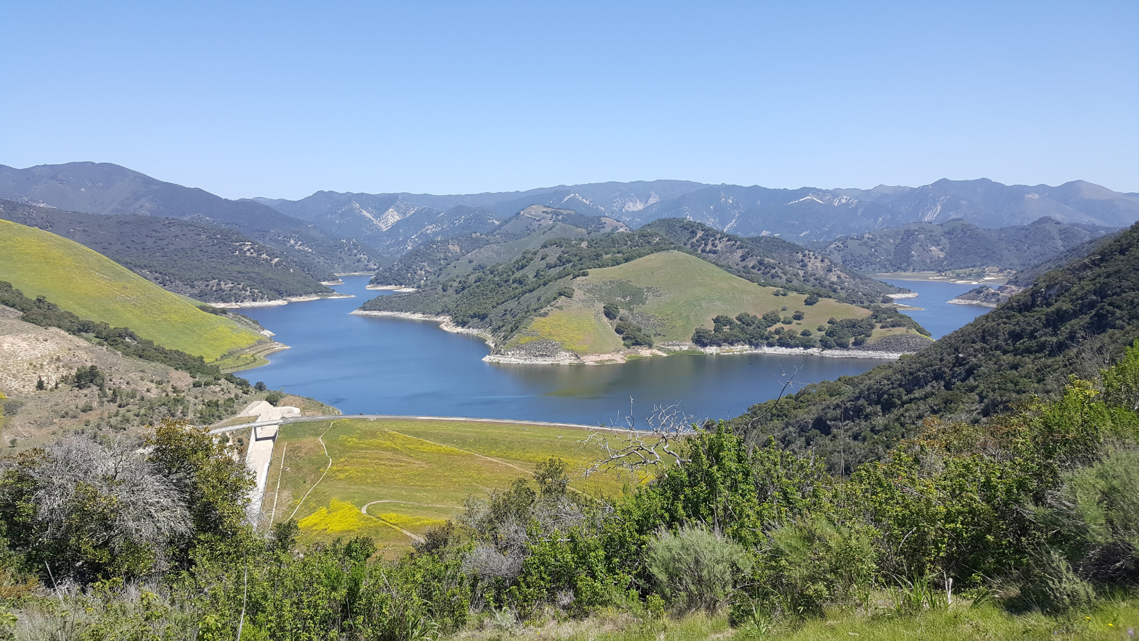A large body of water surrounded by mountains with vegetation and a clear sky above