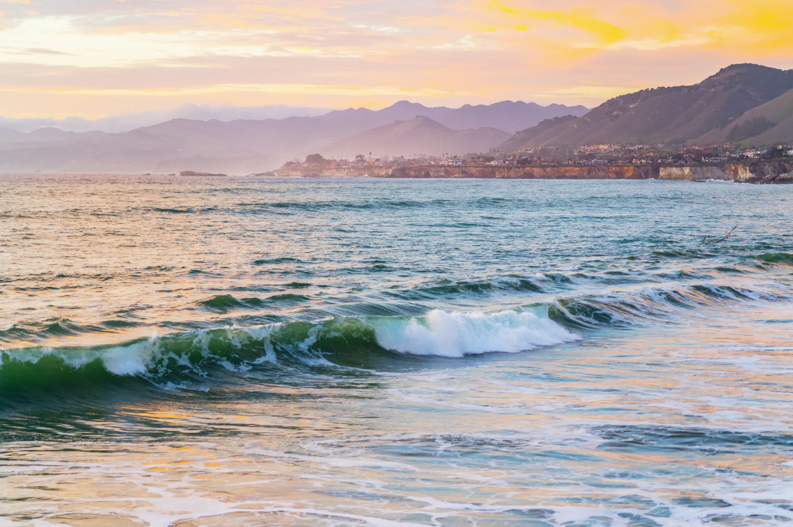 Image of the beach during sunset. The blue/green wave crashing. In the distance, there are large hills and the sky behind it is pink and yellow.