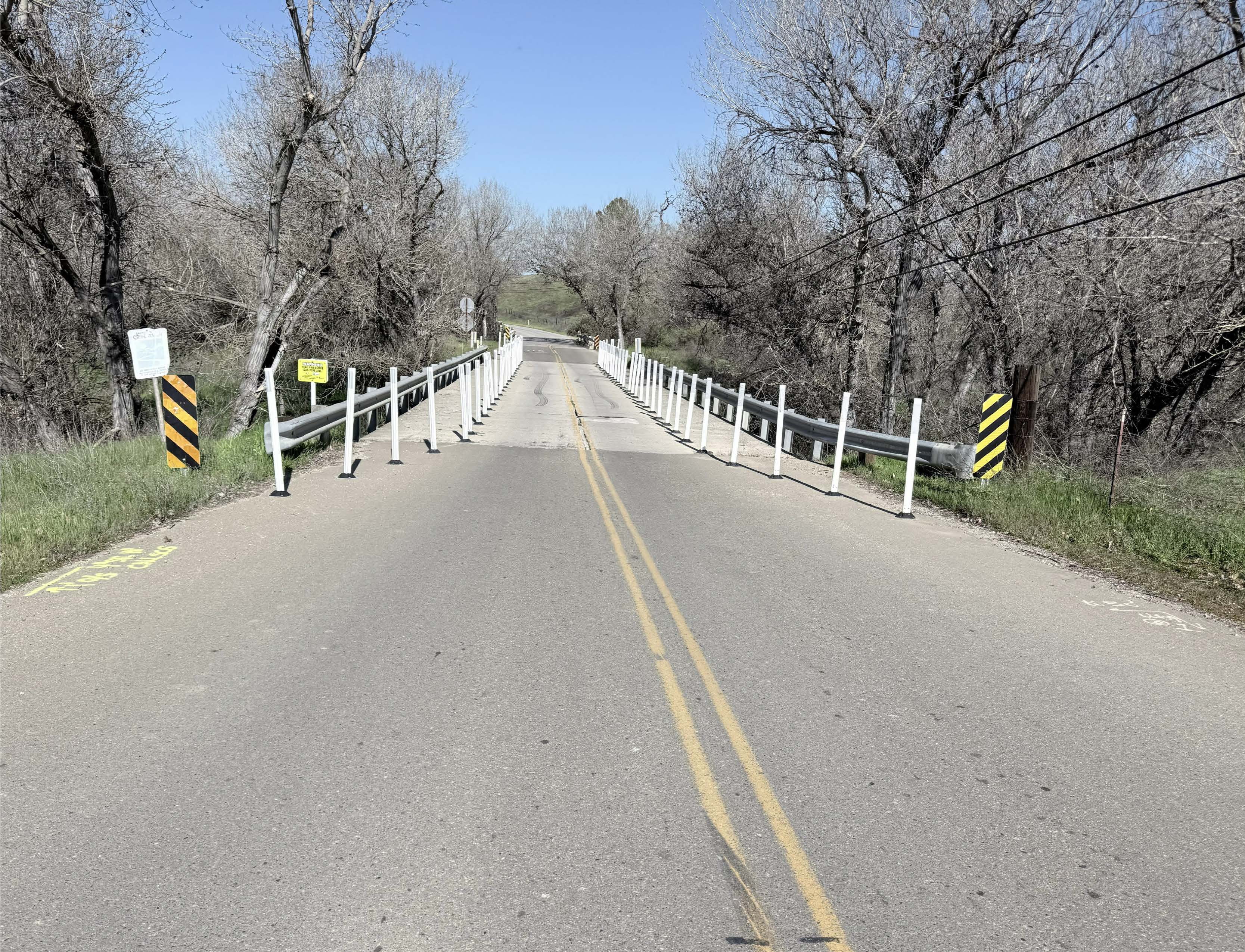 A paved road crosses a narrow bridge with guardrails and white posts, surrounded by leafless trees under a clear blue sky.
