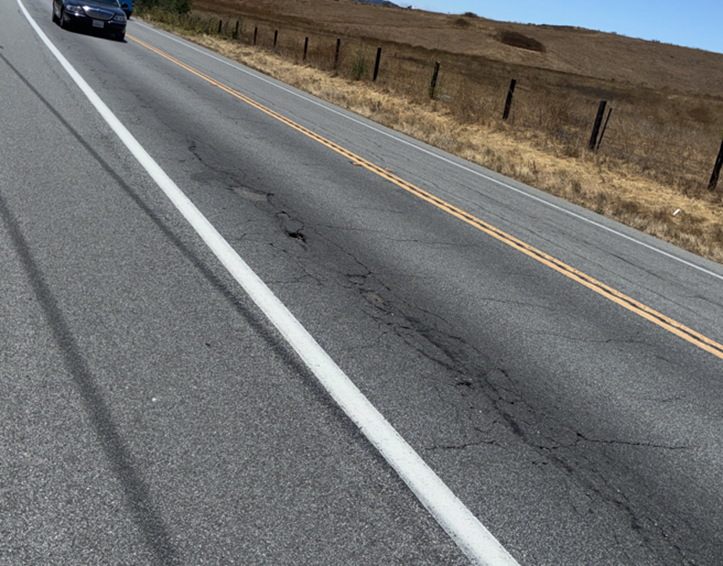 A segment of degraded pavement with a car in the distance and grassy hills beside the road