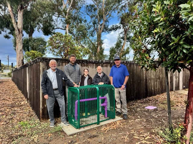 5 people standing and smiling around a pipe that is part of the recycled water project, with a wooden fence behind them, trees and sunny sky with some clouds