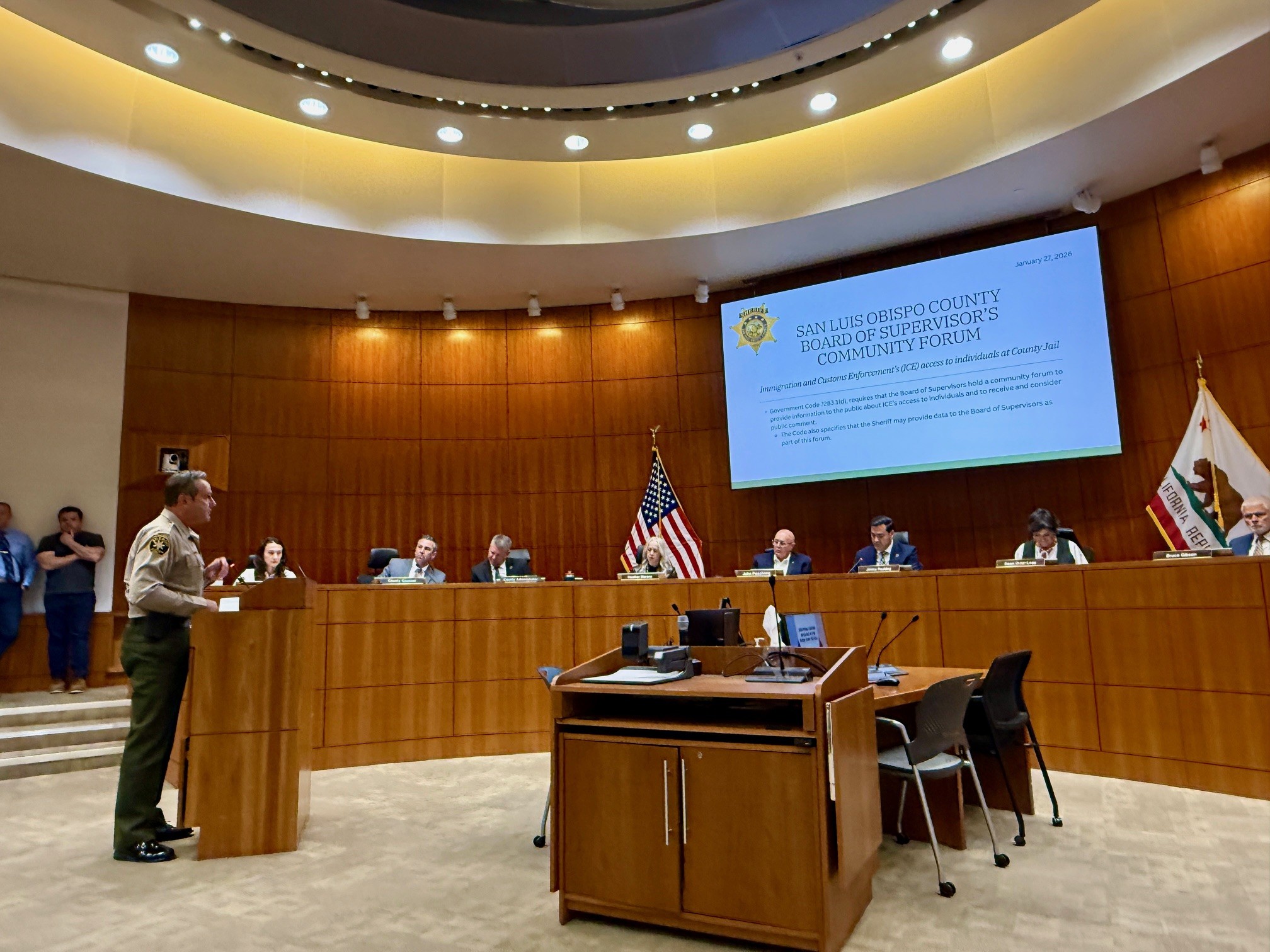 Image of the BOS Chamber. The walls are wood paneled, there is an American flag and a California State flag. In the middle of the wall, there is a video screen with text about the TRUTH forum. 