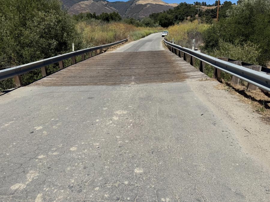 View of a narrow bridge with a wooden deck and metal guardrails, connecting paved road sections with tall grass and hills in the background.