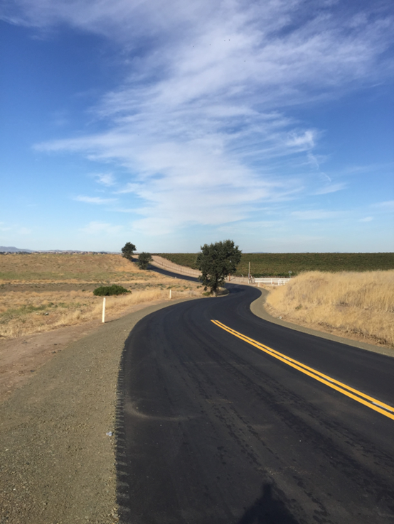 A newly paved rural road curves through dry grassland under a blue sky with scattered clouds.
