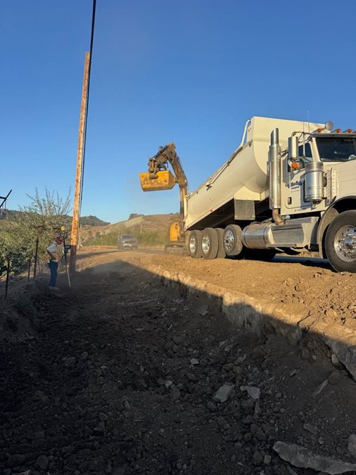 A construction crew works on a dirt road alongside an agricultural field. A large white dump truck and excavator are positioned near a newly installed utility pole, while a worker stands nearby. 