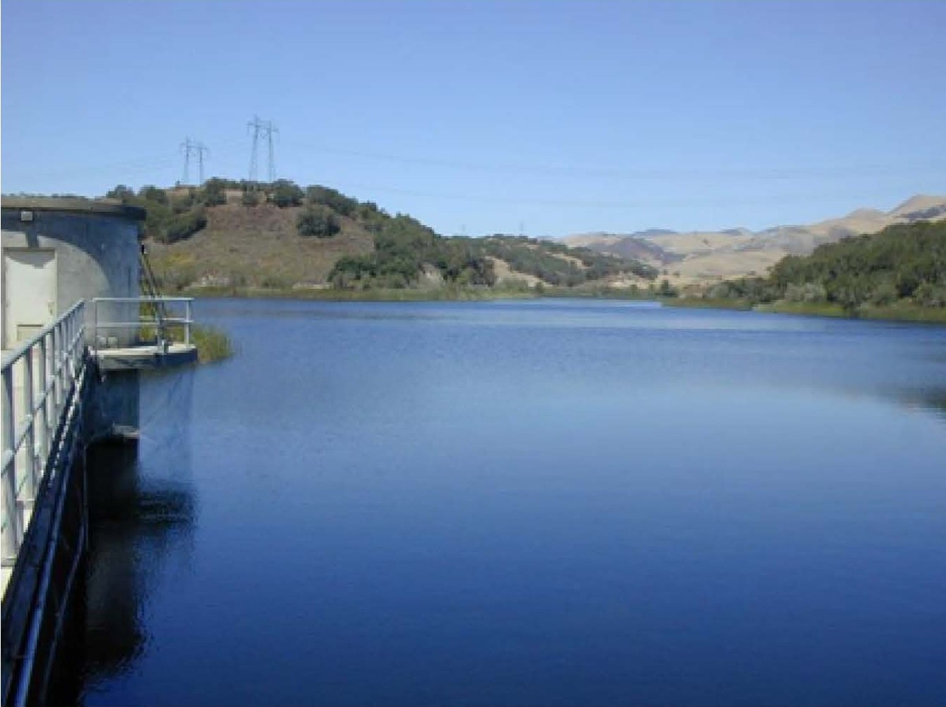 Calm reservoir water with surrounding hills under a clear blue sky.
