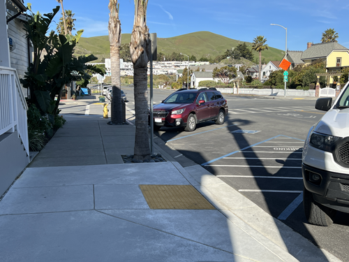 Cars parked on the side of a road near a concrete curb ramp, blue skies and plush green mountains