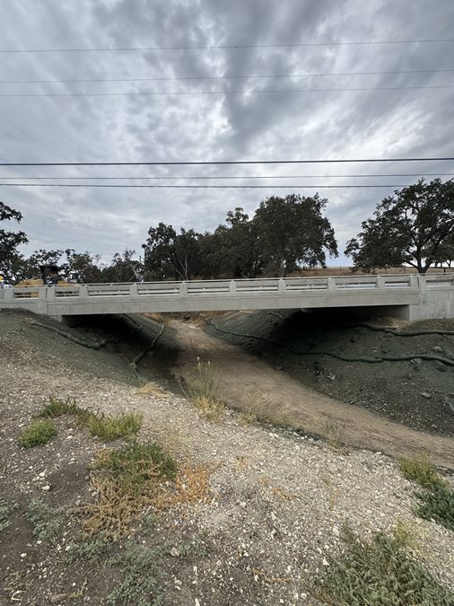 New concrete bridge spanning a dry creek bed in a rural area, surrounded by dirt slopes, erosion-control materials, oak trees, and dry grass. Overhead utility lines cross a cloudy, overcast sky.