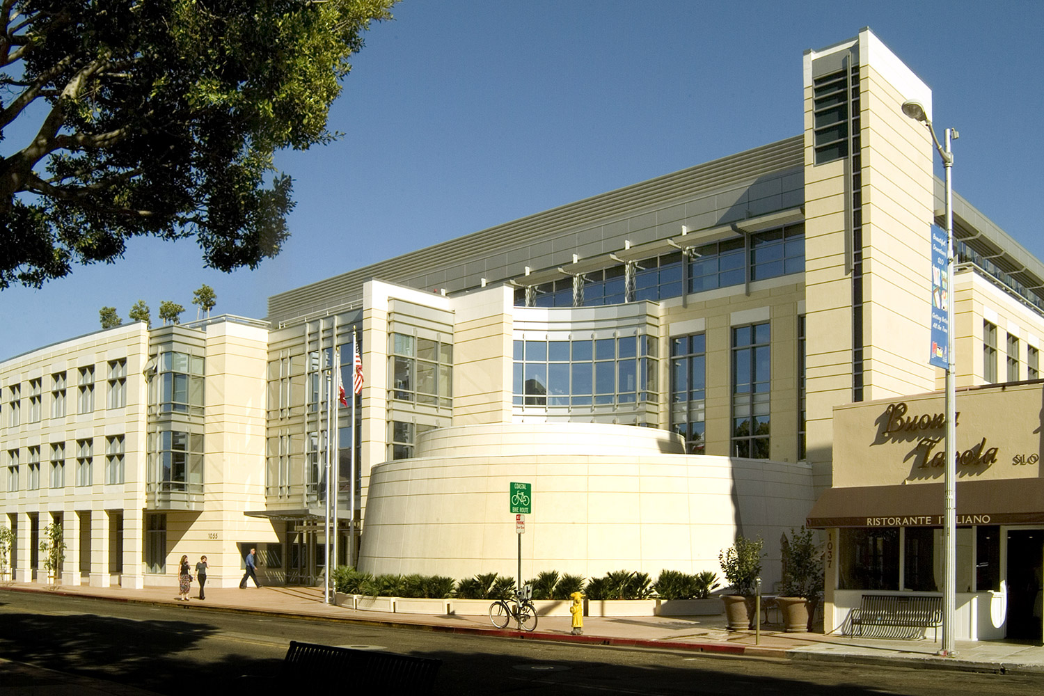 Image of the Katcho Achadjian Government Center which is a cream building with a blue sky. to the left of the image, there are green leaves on tree branches. there is a green sign for the bike lane