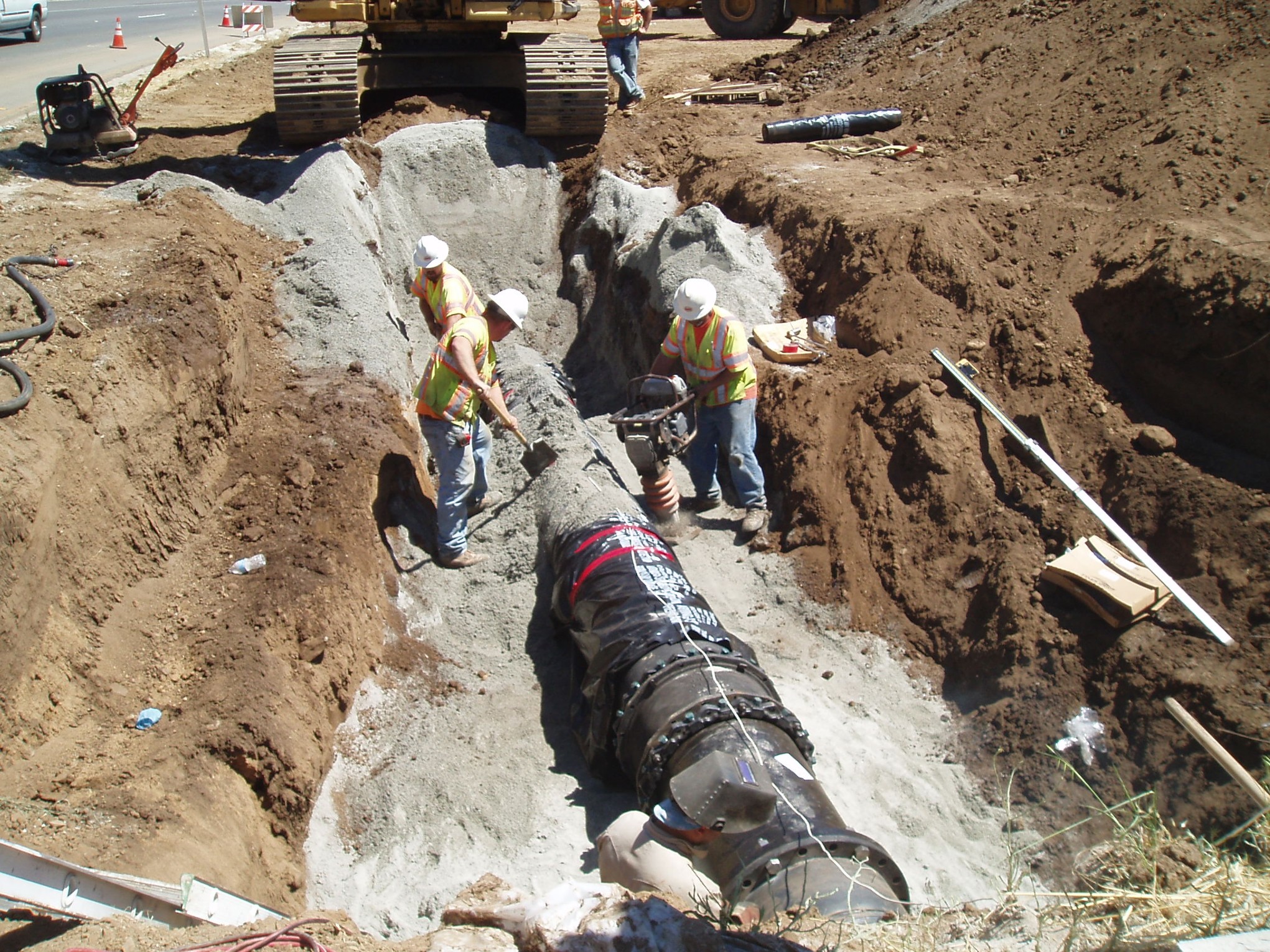 Workers in safety vests and hard hats install a large pipeline in a deep trench, with construction equipment and soil surrounding the work area.