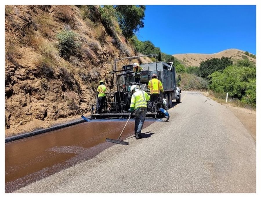 The image shows road workers applying fresh asphalt with a paving machine along the edge of an existing road.