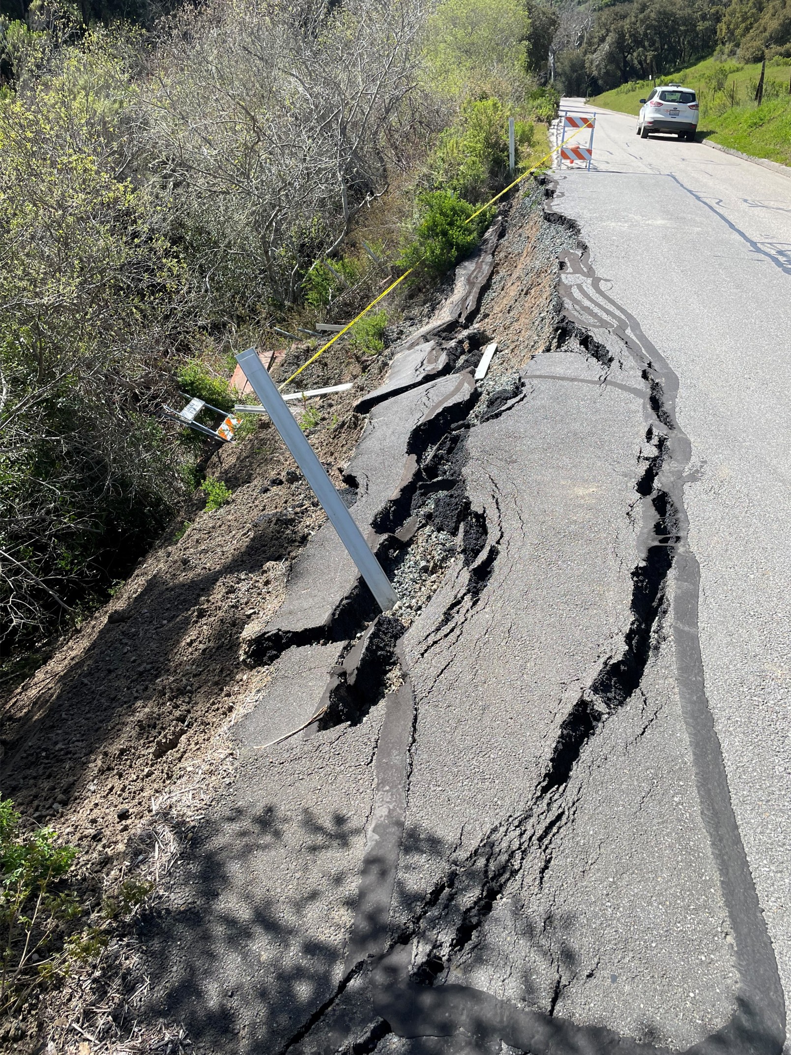 Cracked asphalt on the edge of Perfumo Canyon Road, beside the cliff's edge with stakes and ribbon barriers