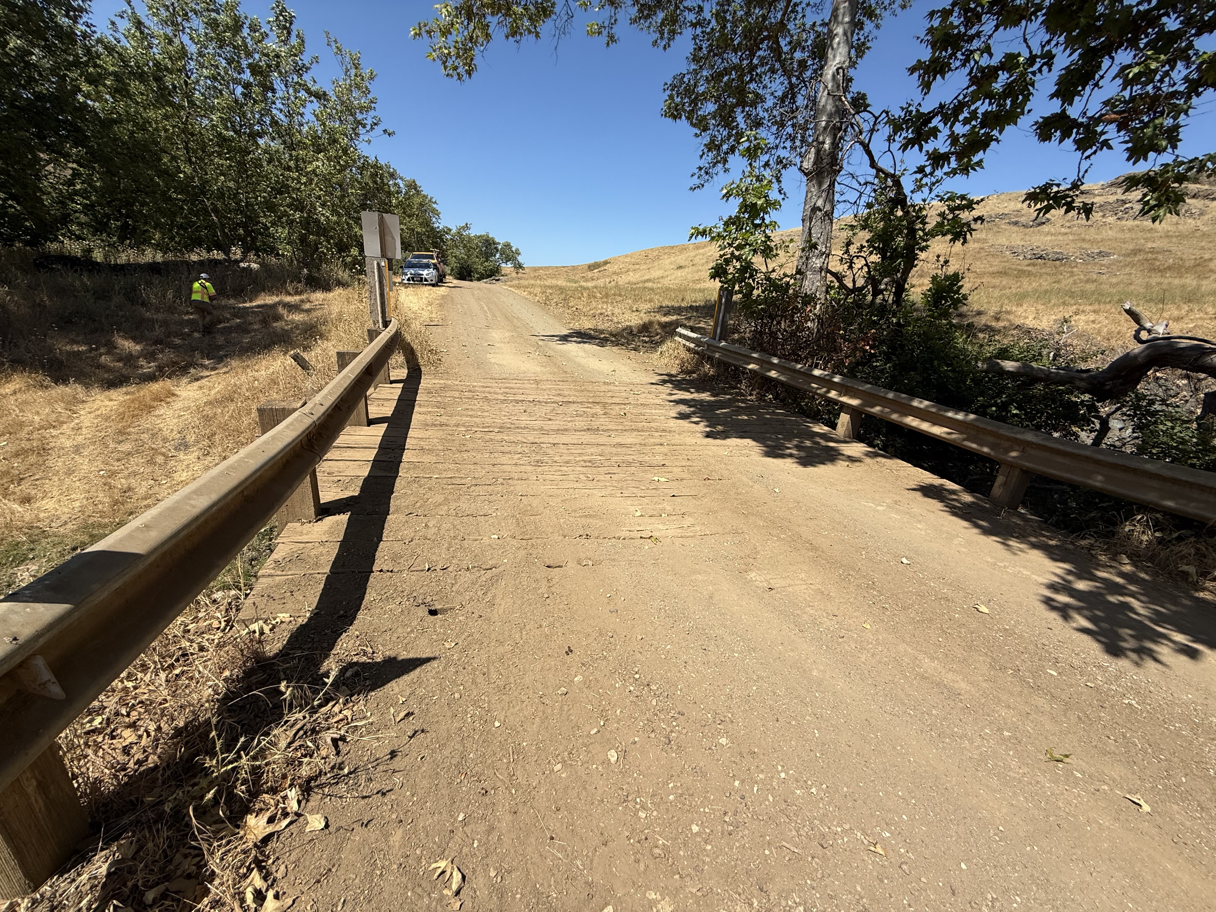 A wooden bridge on a dirt road with foliage, a person, a few vehicles, and blue sky in the background