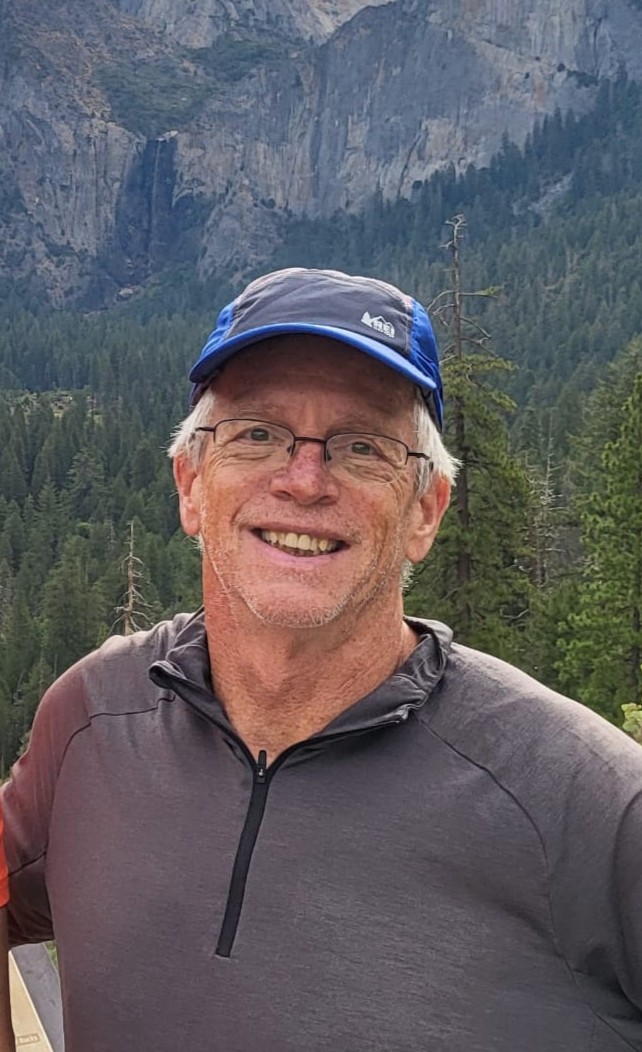 Image of David Cheney wearing a gray shirt, blue hat, and glasses. He stands in front of a scenic mountain with trees below.