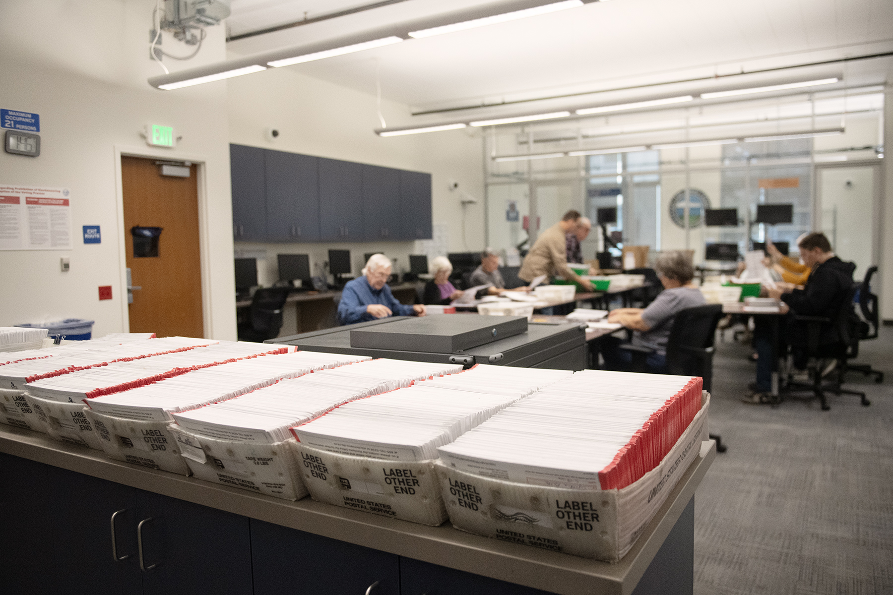 Trays filled with returned ballot envelopes, with election workers at tables in the background