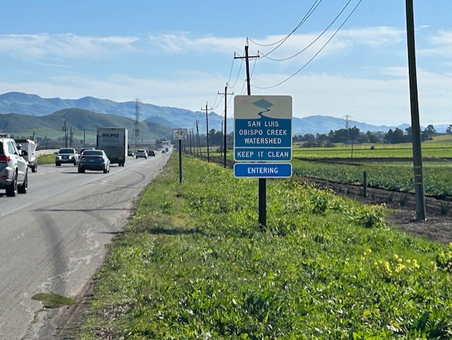 A sign "San Luis Obispo Creek Watershed, Keep It Clean" is in the shoulder of a busy two-lane road, beside telephone lines and green farmland with green hills, mountains, and clouds in the distance 