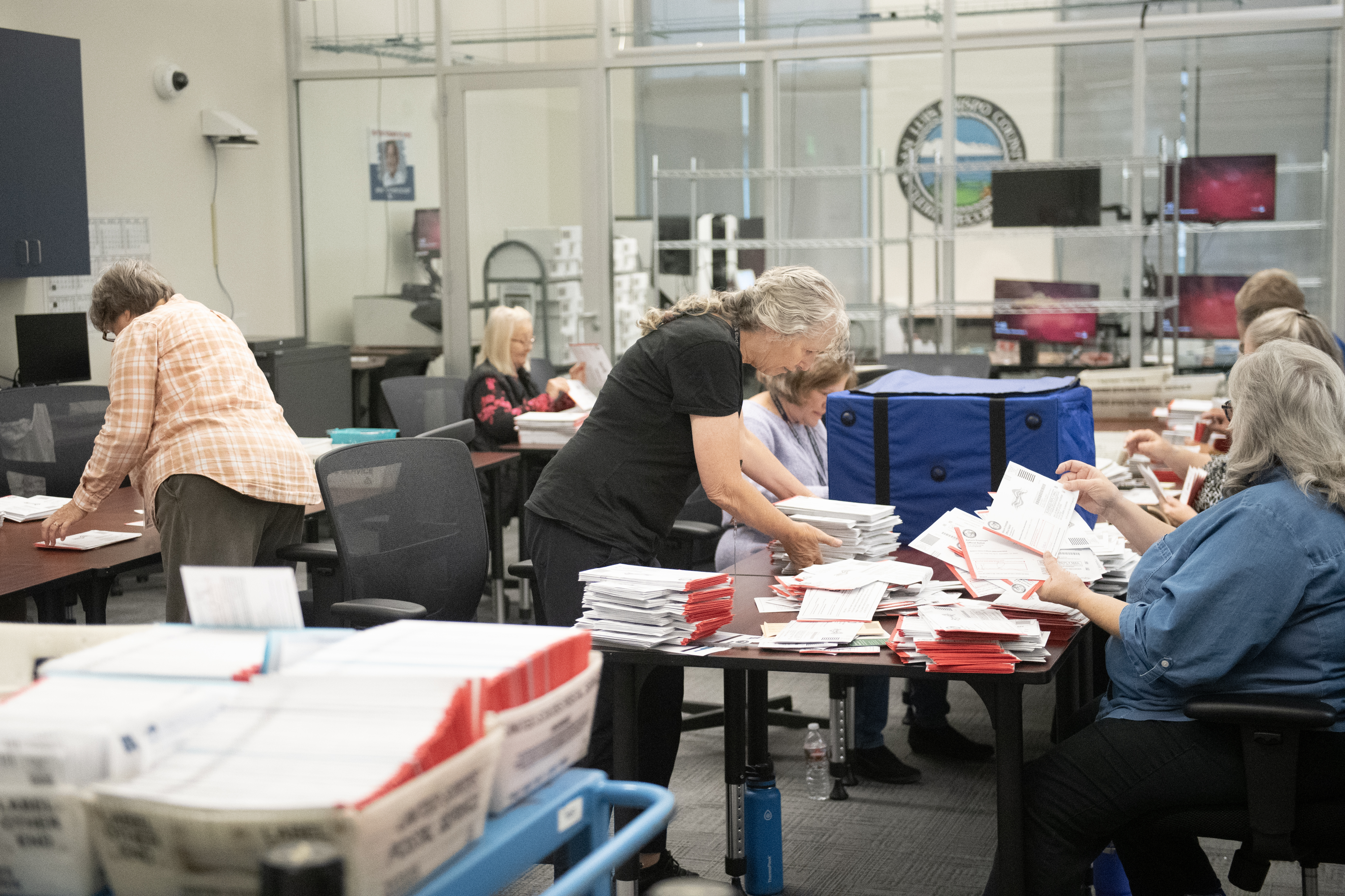 A woman standing at a table with piles of ballots while others sit and work around her