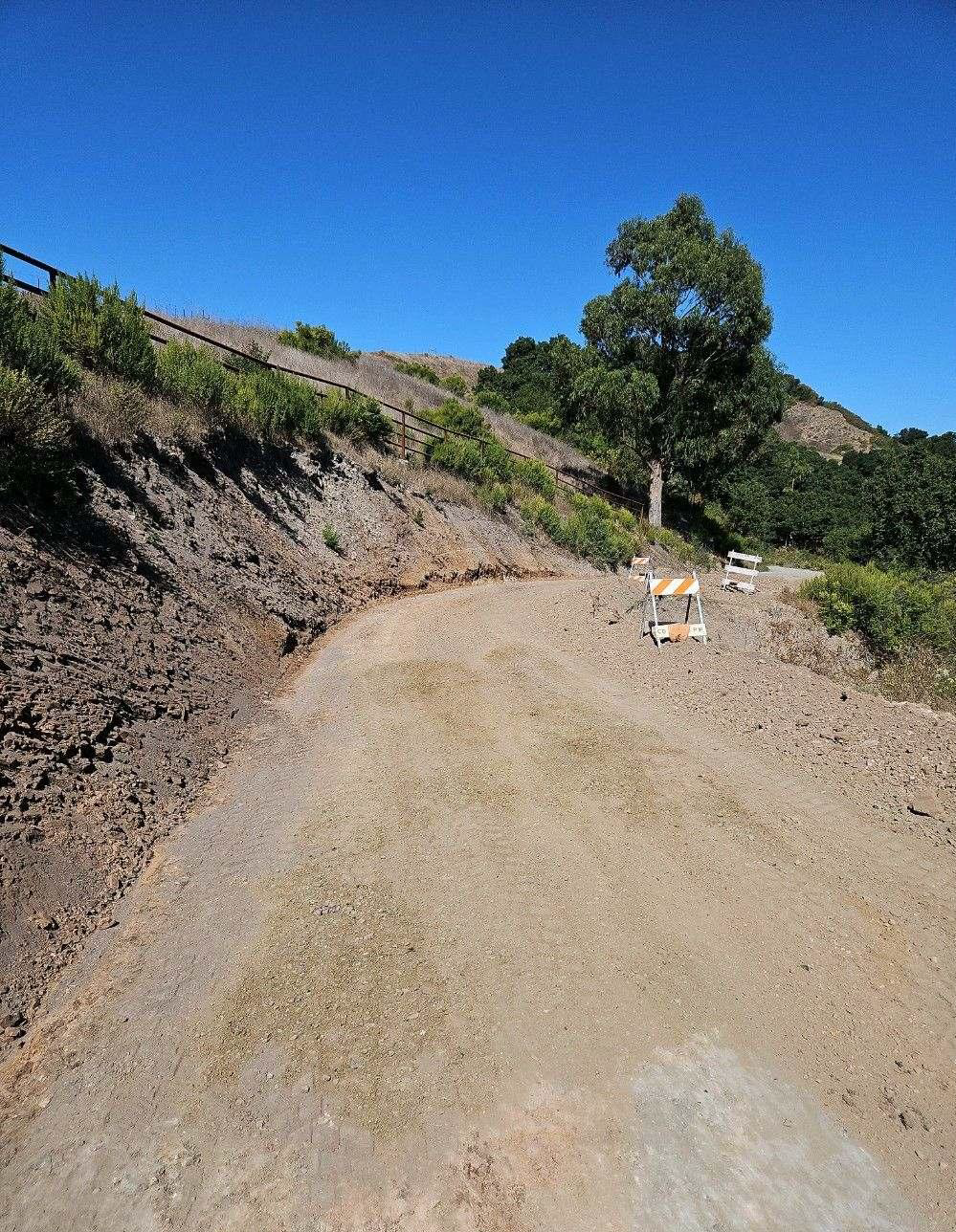 A dirt road curving beside on a hillside with a metal fence, construction signs, brush and trees beneath the clear sky