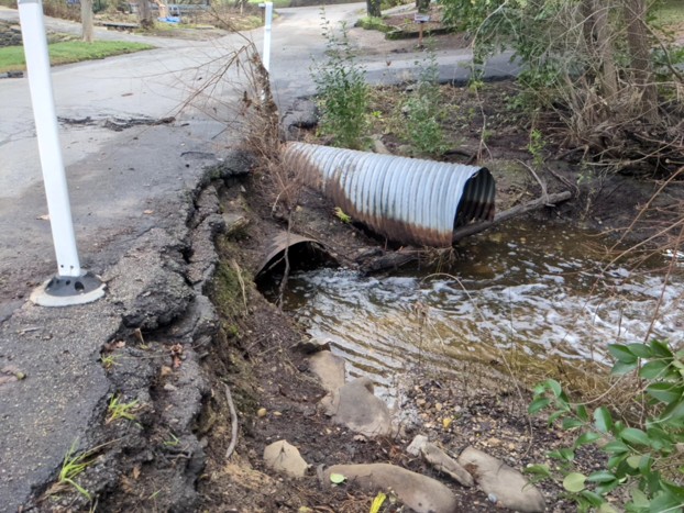 Damaged roadway with an exposed drainage pipe over a flowing stream.