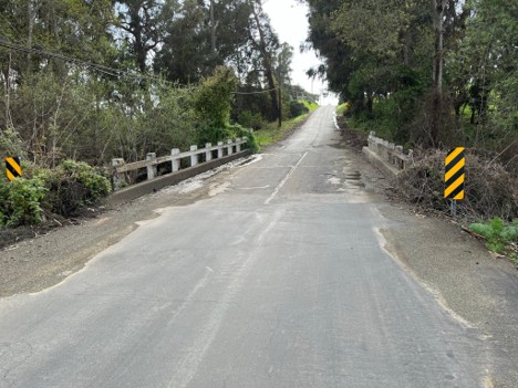 A narrow paved road crosses a small bridge with concrete guardrails and black-and-yellow hazard markers, surrounded by dense trees and vegetation.