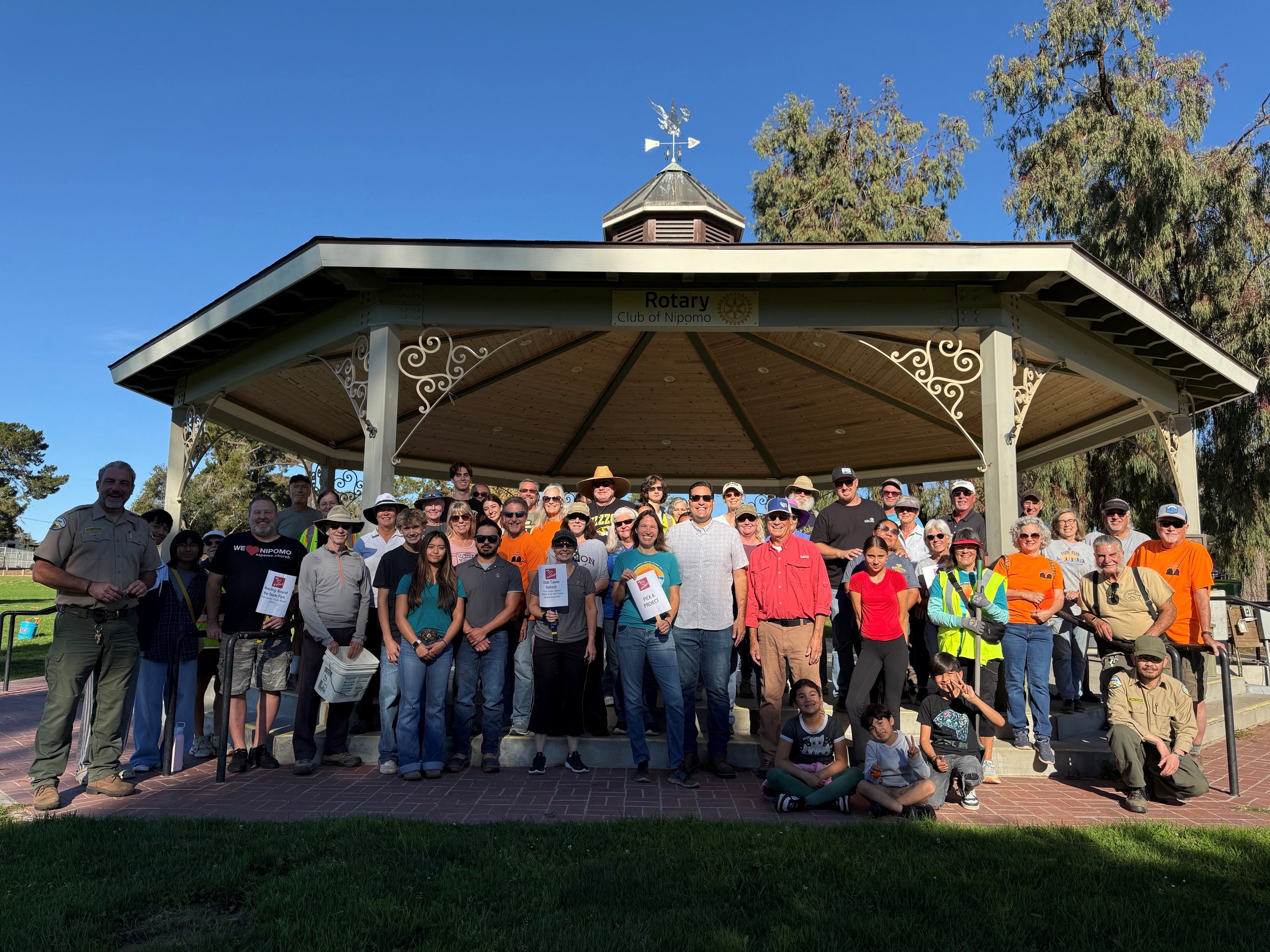 Image of a clear blue sky. Standing under a gazebo that says Rotary Club of Nipomo. Some people have signs that say state volunteer jobs.