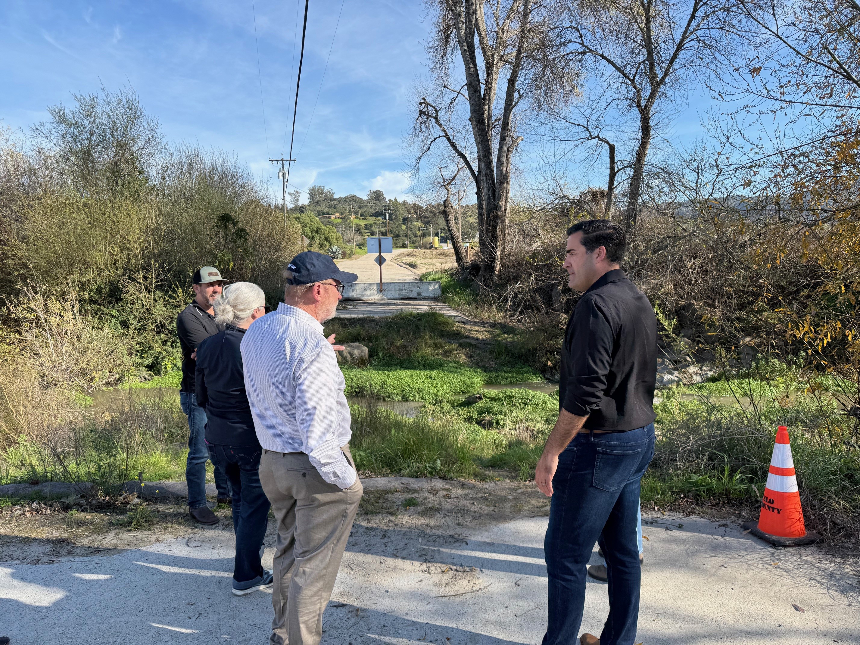 Image of Supervisor Jimmy Paulding standing with Senator Laird. They are outside looking at road damage. In the foreground are two other people and beyond them is bushes. There is an orange cone 