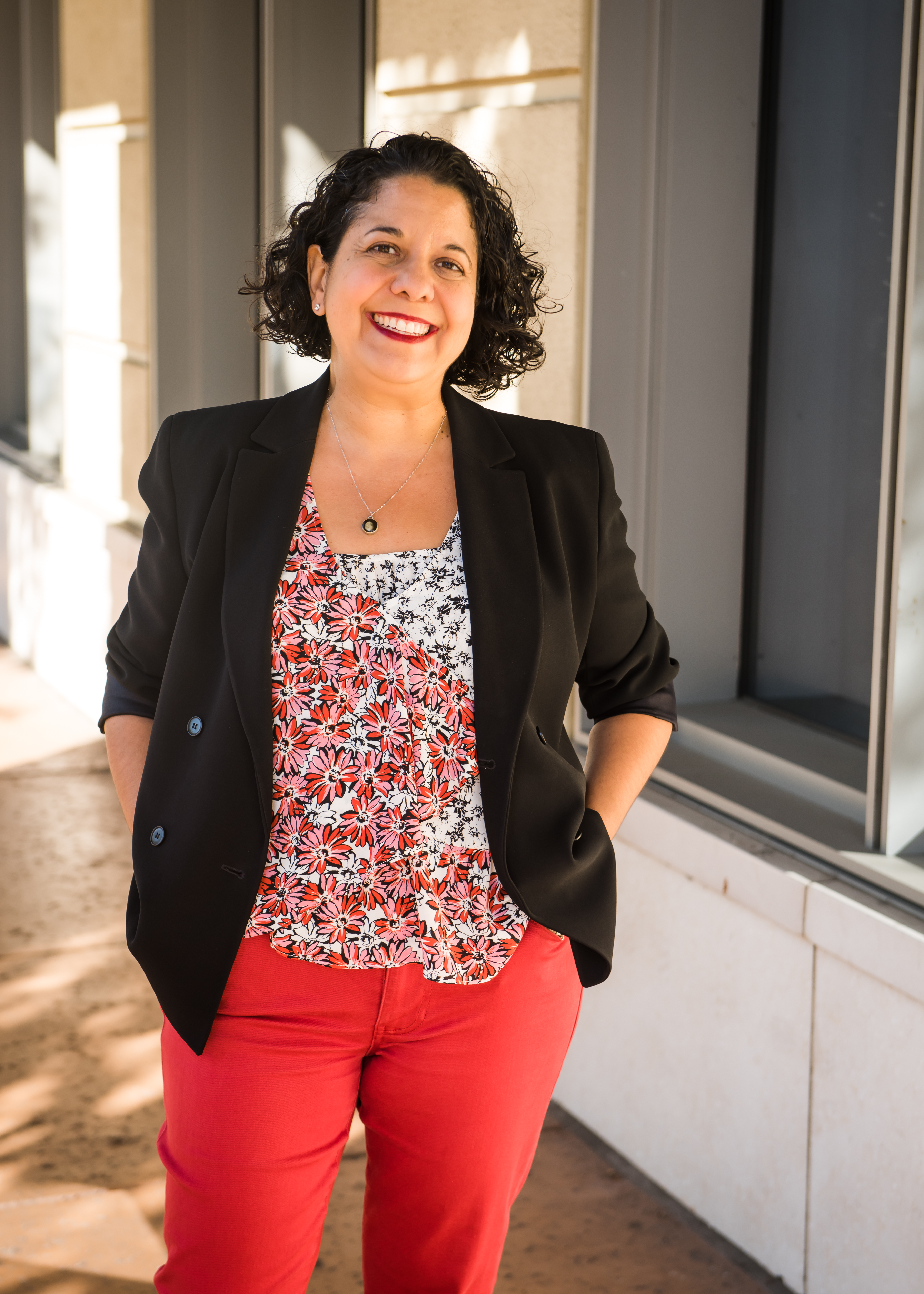 Photo of Erica standing outside. She has red pants and a flower top paired with a black blazer.