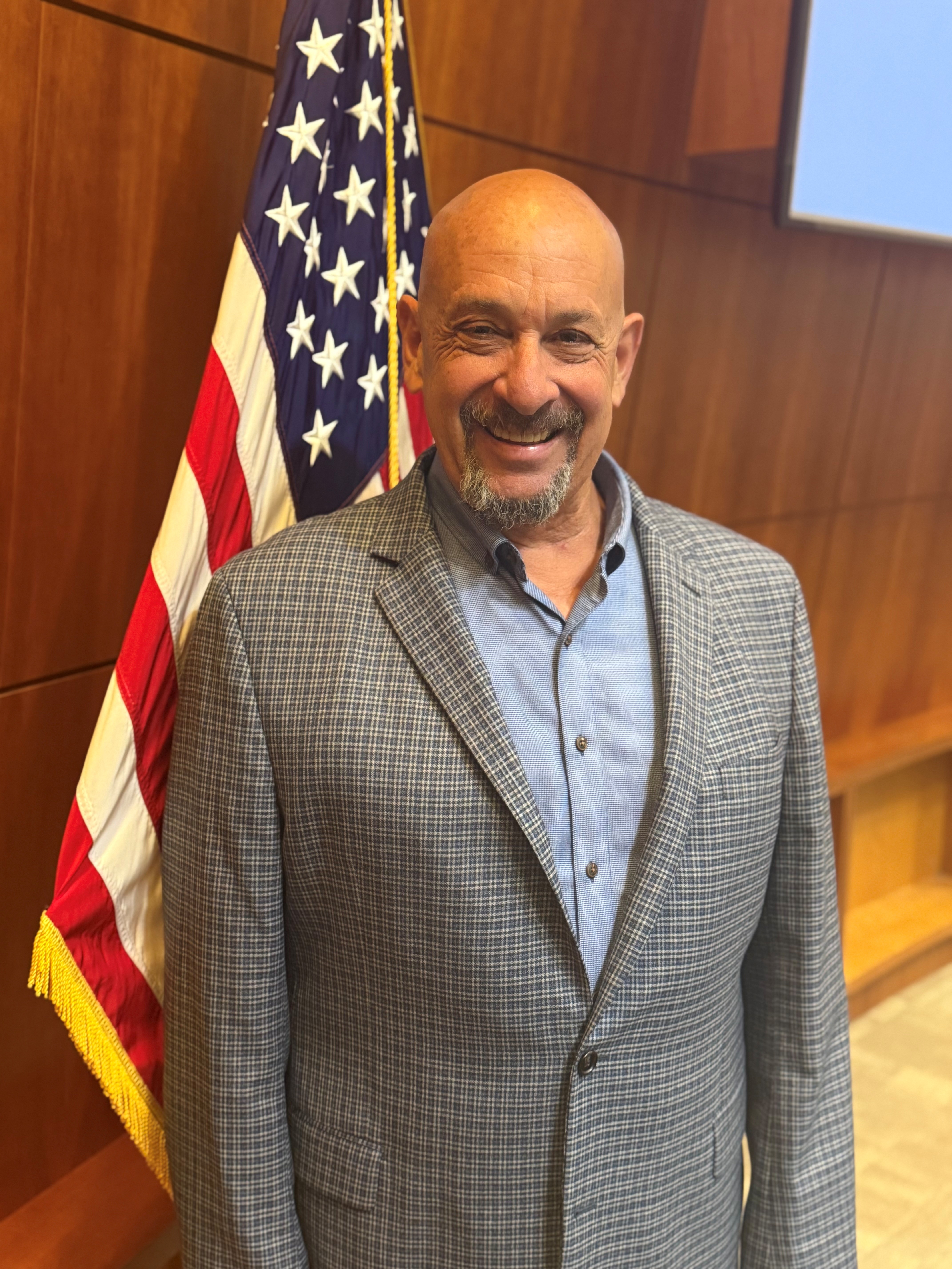 Steve Lieberman standing in front of the American flag. He is wearing a blue shirt with a gray jacket.